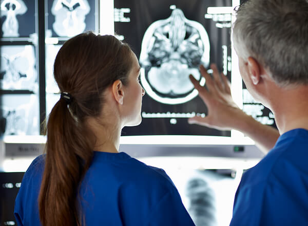 Two medical professionals in blue scrubs examine a brain scan displayed on a monitor, with one pointing at details on the image.