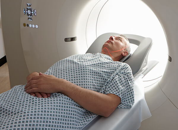 A man in a hospital gown lies on his back inside a CT scanner, preparing for a medical imaging procedure.