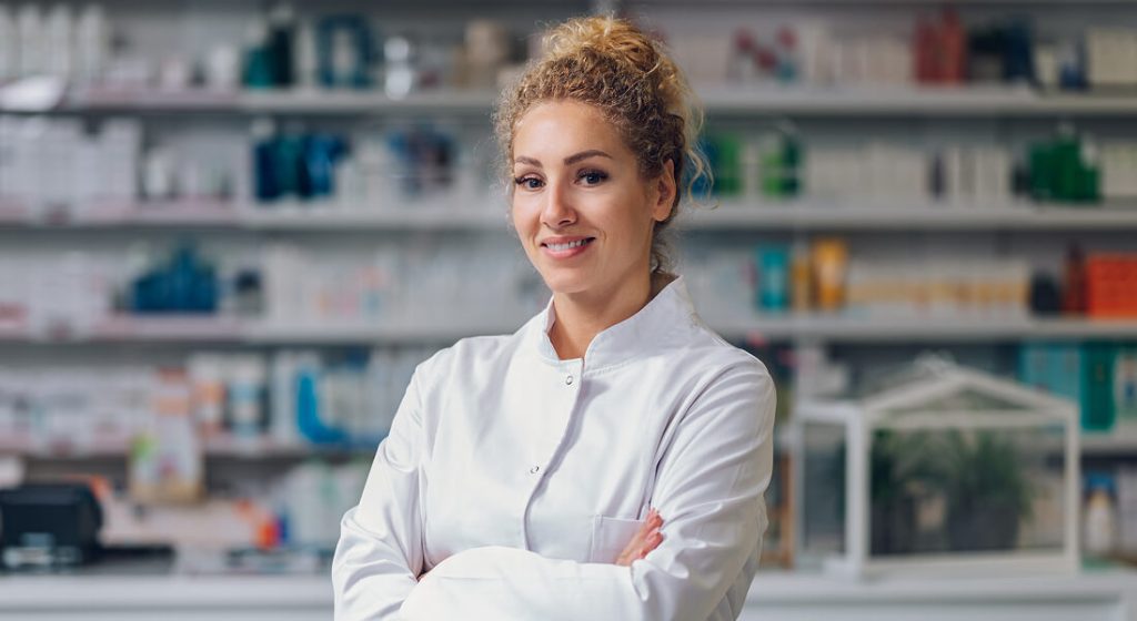 A woman in a white lab coat stands with arms crossed, smiling, in front of shelves stocked with pharmaceutical products in a pharmacy.