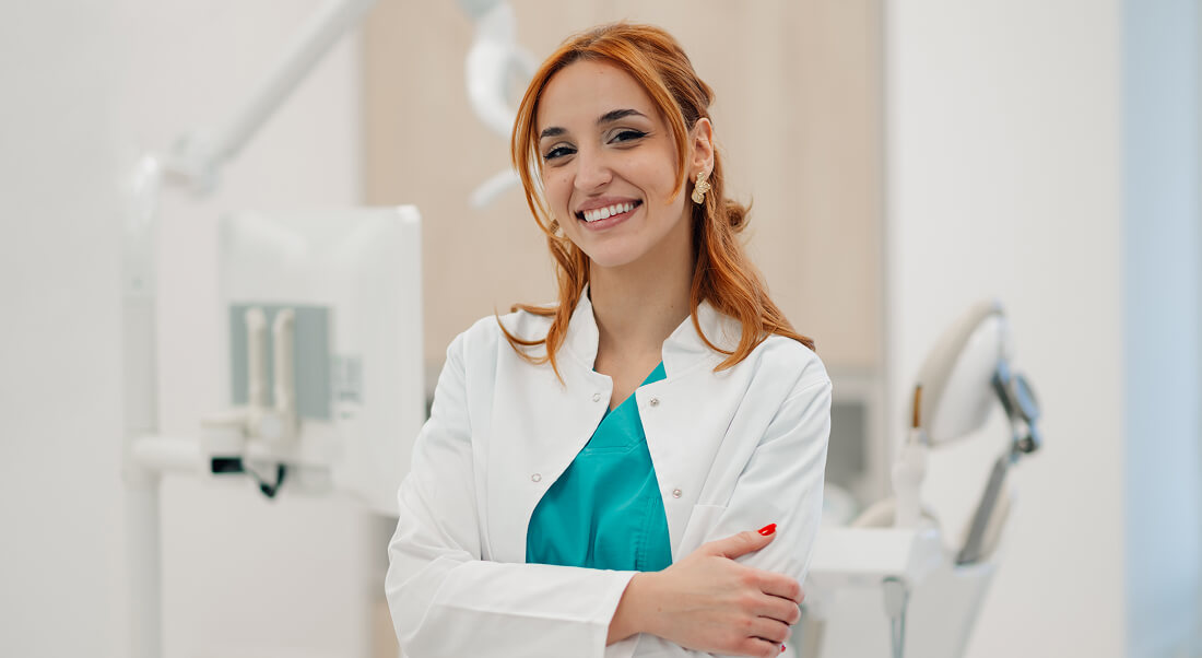 A woman in a white lab coat stands smiling with arms crossed in a dental office, with dental equipment visible in the background.