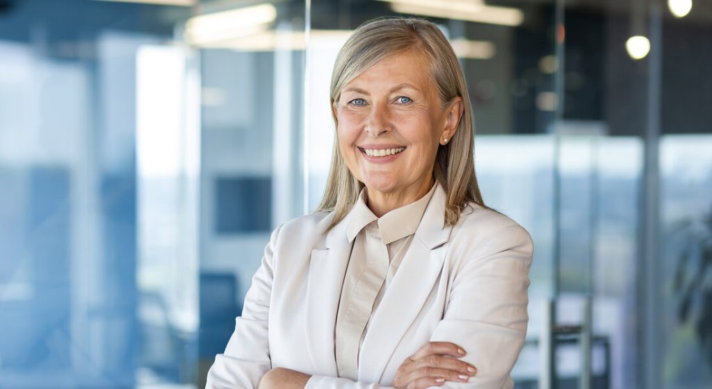 Smiling older woman in a light-colored suit stands with arms crossed in a modern office with glass walls.