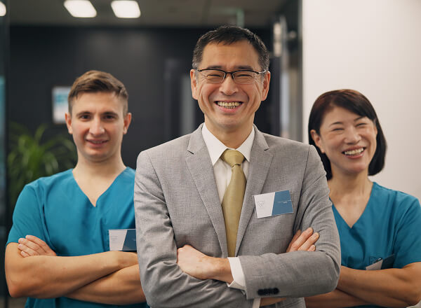 Three people pose indoors; a man in a suit stands in front, smiling, with two people in blue scrubs smiling behind him.