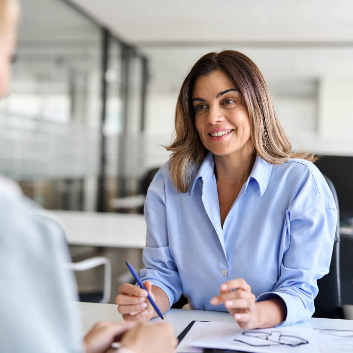 Woman in a blue shirt sits at a desk, holding a pen and smiling, during a conversation in a modern office setting.