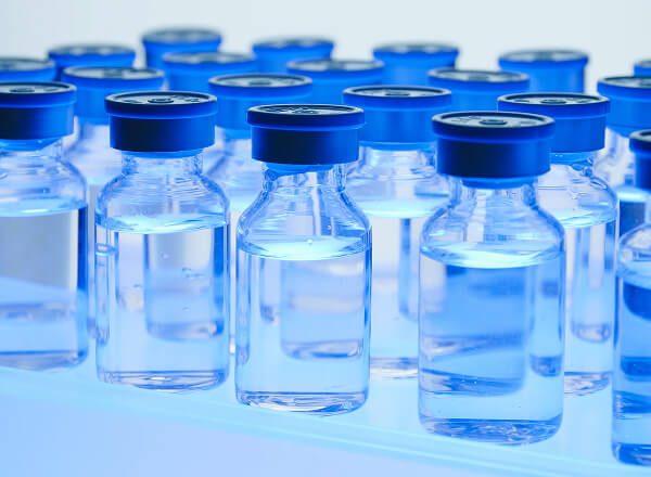 Clear glass vials with blue caps are arranged in rows on a reflective surface, containing a transparent liquid.
