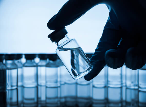 A gloved hand holds a small glass vial filled with clear liquid; several similar vials are lined up in the background.