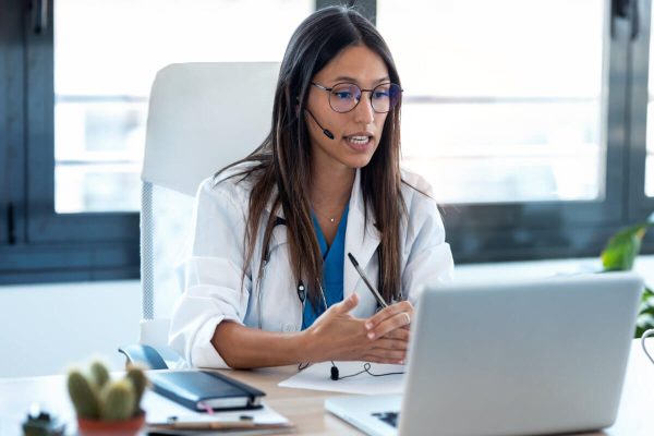 A doctor wearing a headset and white coat sits at a desk, speaking during a video call on a laptop, with documents and a stethoscope in view.
