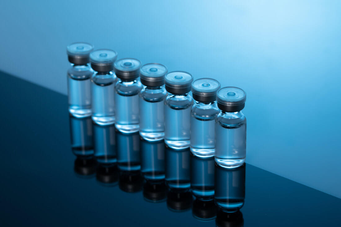Seven clear glass vials with silver caps are lined up on a reflective black and blue surface against a blue gradient background.
