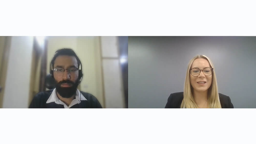 A man and a woman are participating in a virtual video call. The man has a beard and wears headphones. The woman has long hair and glasses. Both appear to be indoors.