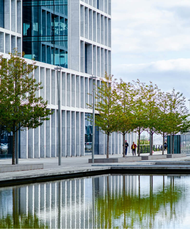 Modern office building with reflective glass facade, lined with trees beside a water feature, and two people walking in the distance.