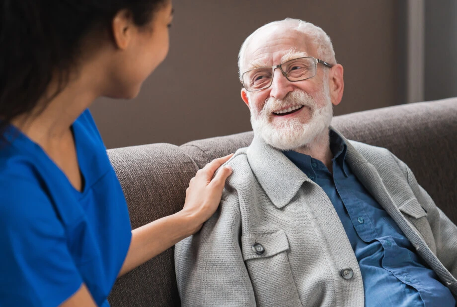 A smiling older man with glasses sitting on a couch, looking at a person in blue scrubs who is touching his shoulder.