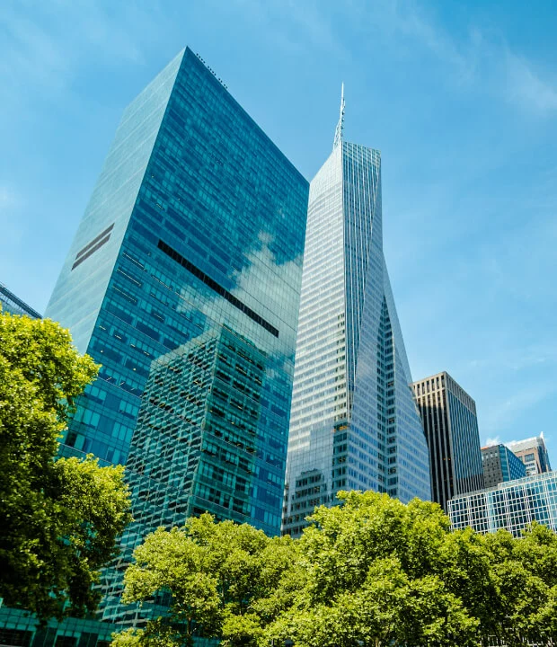 Tall modern skyscrapers with glass facades and reflections, surrounded by green trees under a clear blue sky.