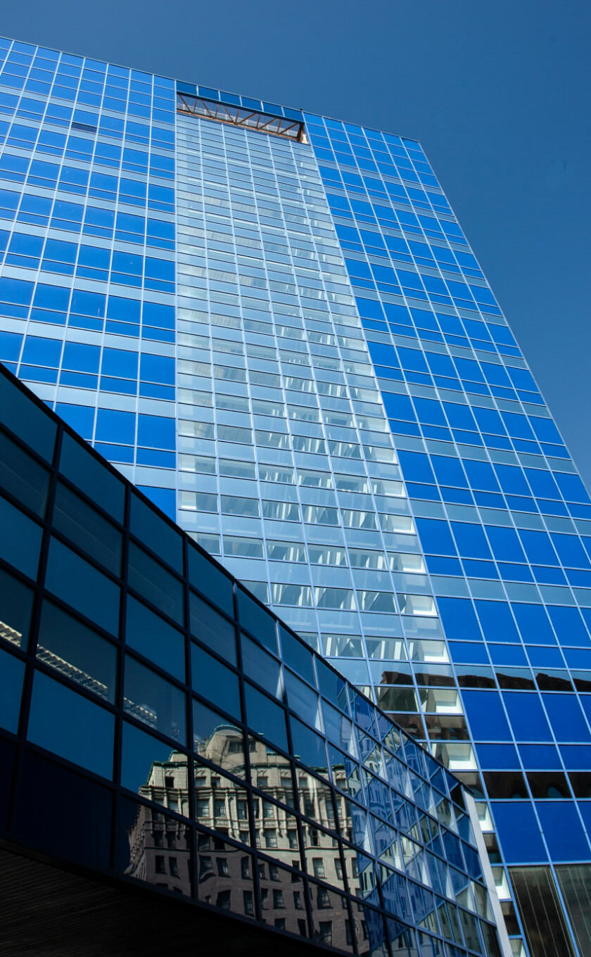 A tall, blue glass skyscraper reflecting sunlight. Another building is visible in the reflection on the lower section. Clear blue sky in the background.