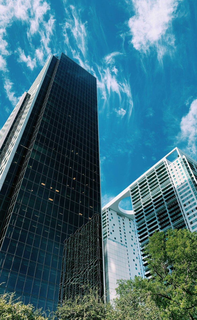 Tall glass skyscraper and a modern white building against a blue sky with clouds, surrounded by green trees.