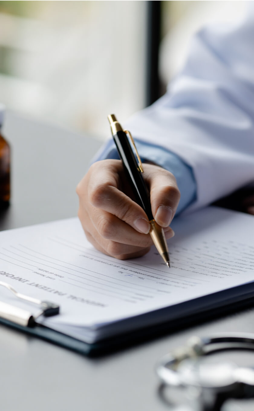 Close-up of a person writing on a clipboard with a pen. A stethoscope and a small medicine bottle are visible on the table.
