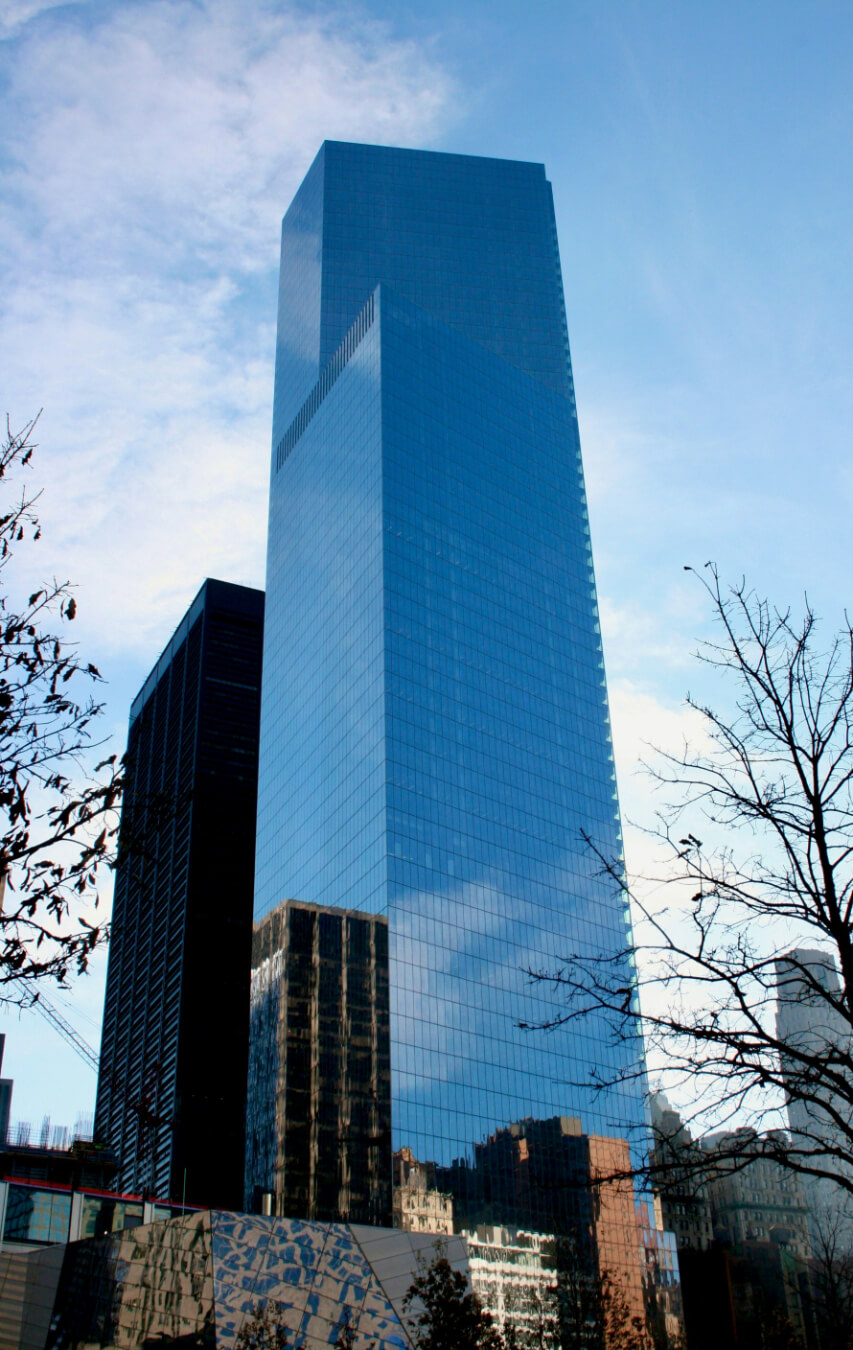 A tall, reflective glass skyscraper rises against a blue sky, with trees and smaller buildings in the foreground.