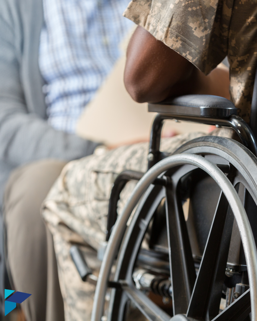 Person in military uniform seated in a wheelchair, holding a folder, with another person sitting nearby in the background.