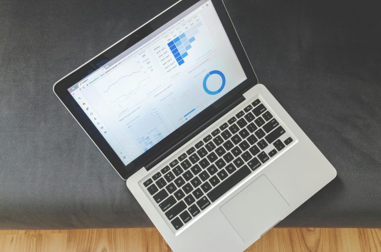 Open laptop displaying graphs and charts on a data analytics dashboard, situated on a dark grey surface with a wooden floor partially visible.