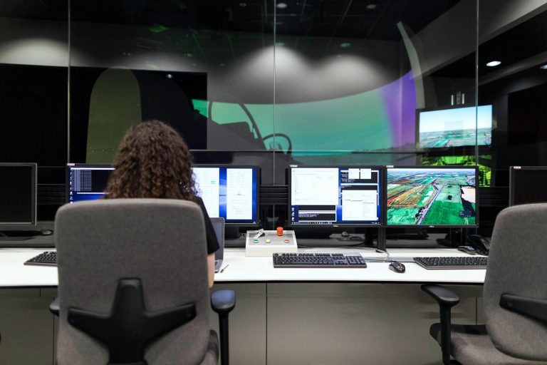 A person sits at a desk with multiple computer monitors displaying data and images in a control room.