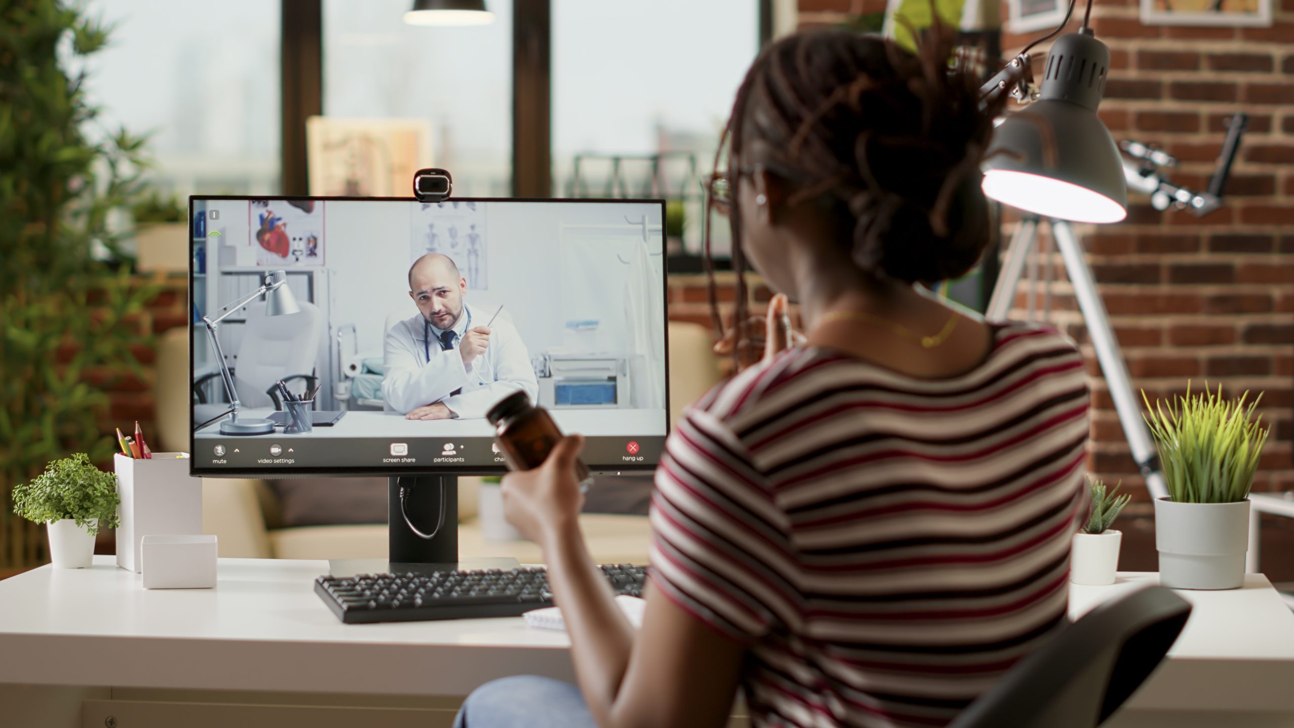 A woman holds a medication bottle while having a video call with a doctor, who appears on her computer screen in a medical office setting.
