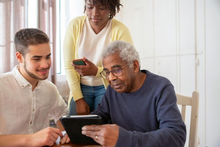 Three people, two men and one woman, look at a tablet together at a table in a bright room. The older man is seated, while the younger man and woman stand beside him.