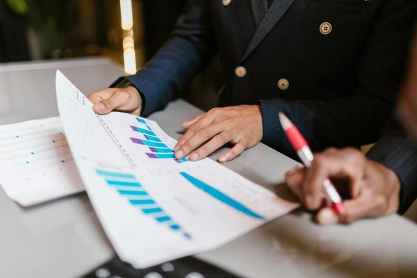 Two people in business attire review and discuss printed charts and graphs at a desk, with one person holding a pen and the other pointing to the data.