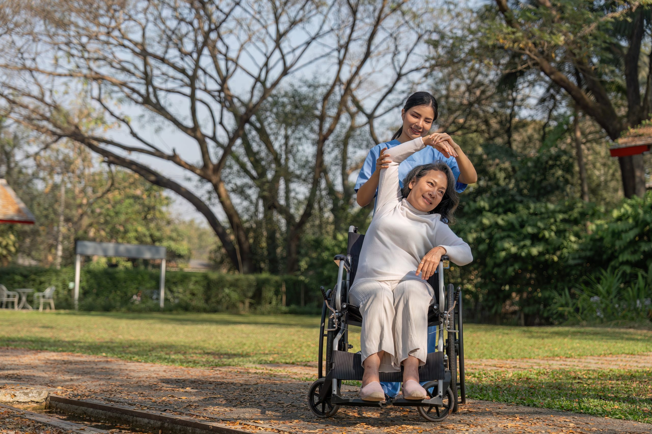 A caregiver assists an older woman in a wheelchair with arm exercises in an outdoor park setting.