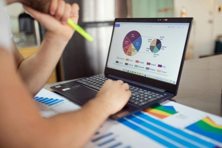 Person analyzing colorful pie and bar charts on a laptop, with additional printed graphs and a highlighter on the table.