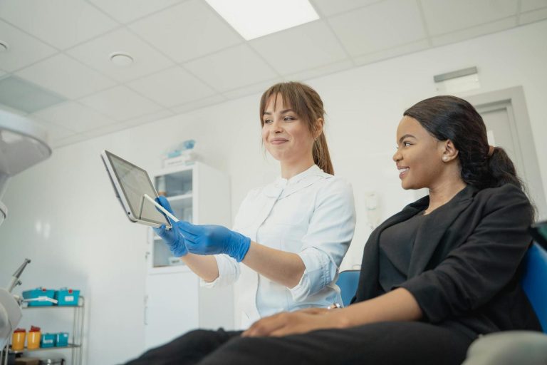 A healthcare professional wearing gloves shows a tablet to a seated patient in a medical office.