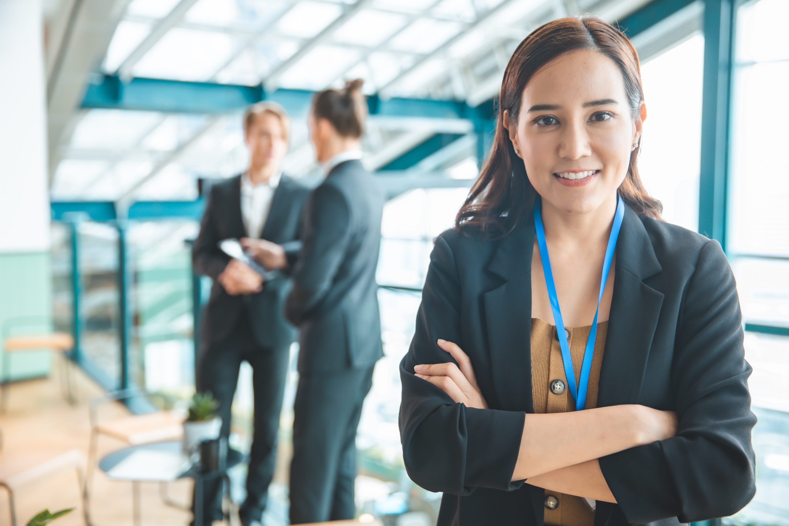 A woman in business attire stands with arms crossed and smiles at the camera; two people converse in the background in a modern office setting.