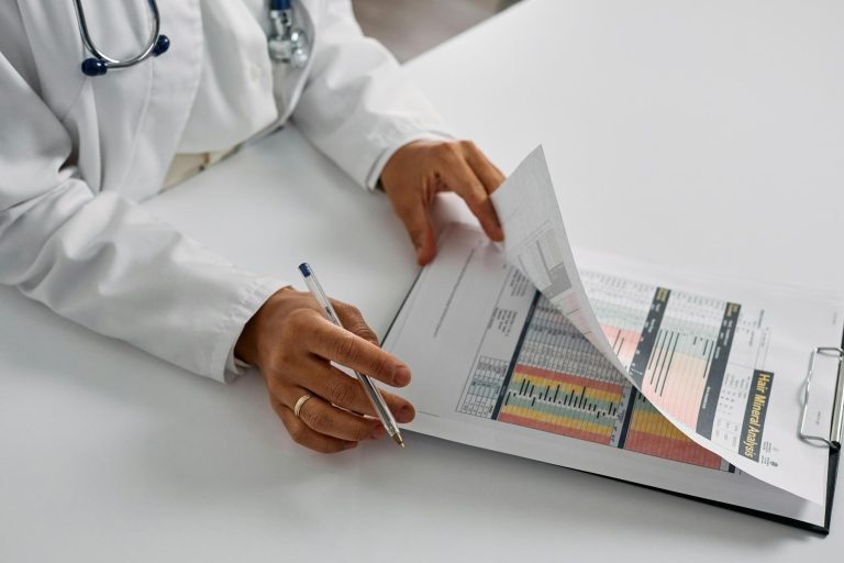 A person in a white coat reviews printed charts and data sheets on a clipboard, holding a pen and flipping through the pages at a desk.