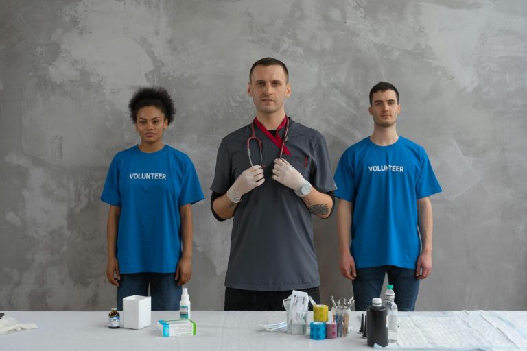Three people stand behind a table with medical supplies; two wear blue "Volunteer" shirts, and the person in the center wears medical scrubs and gloves with a stethoscope around his neck.