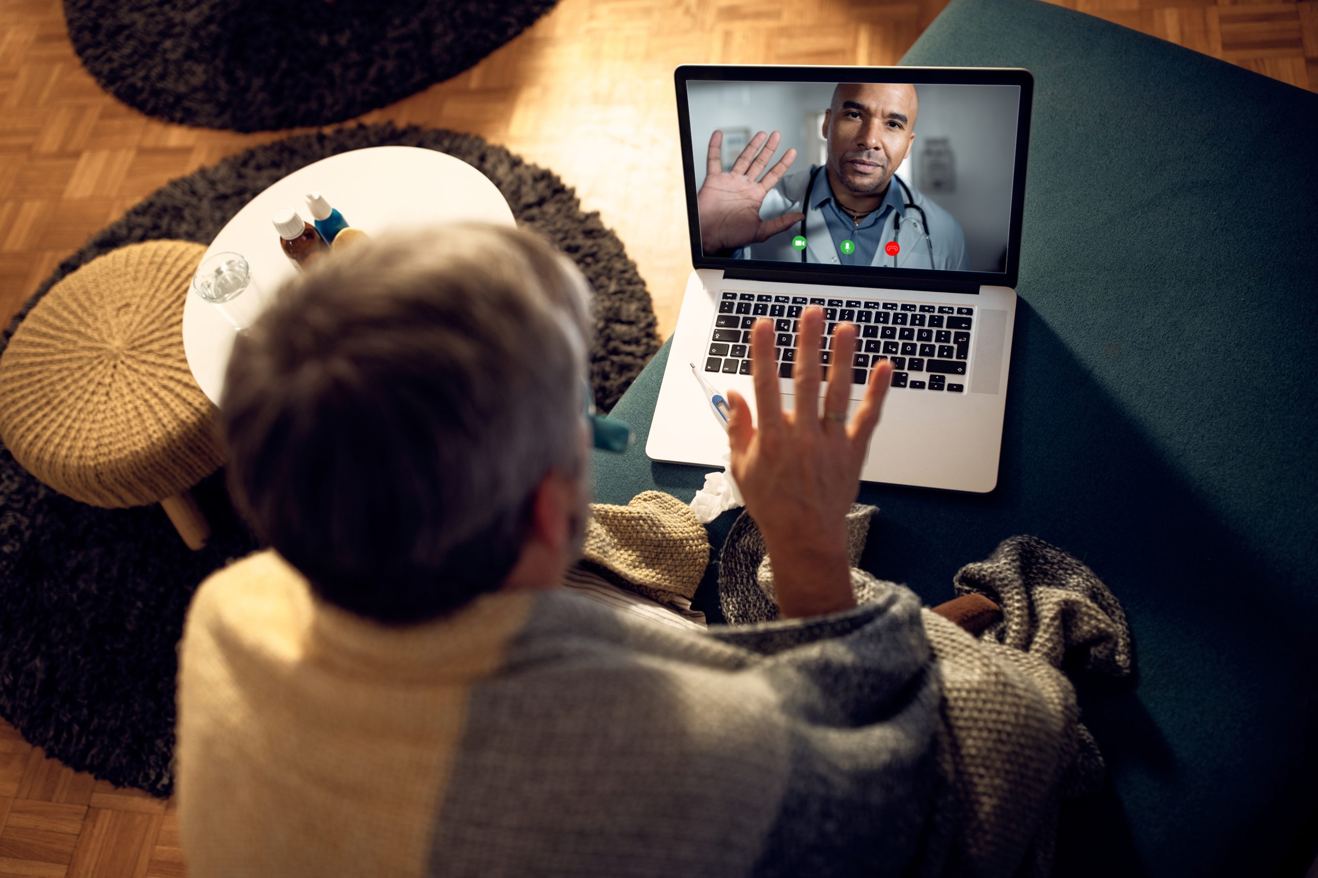 Person sitting on a couch, wrapped in a blanket, having a video call with a doctor on a laptop, both raising a hand in greeting.