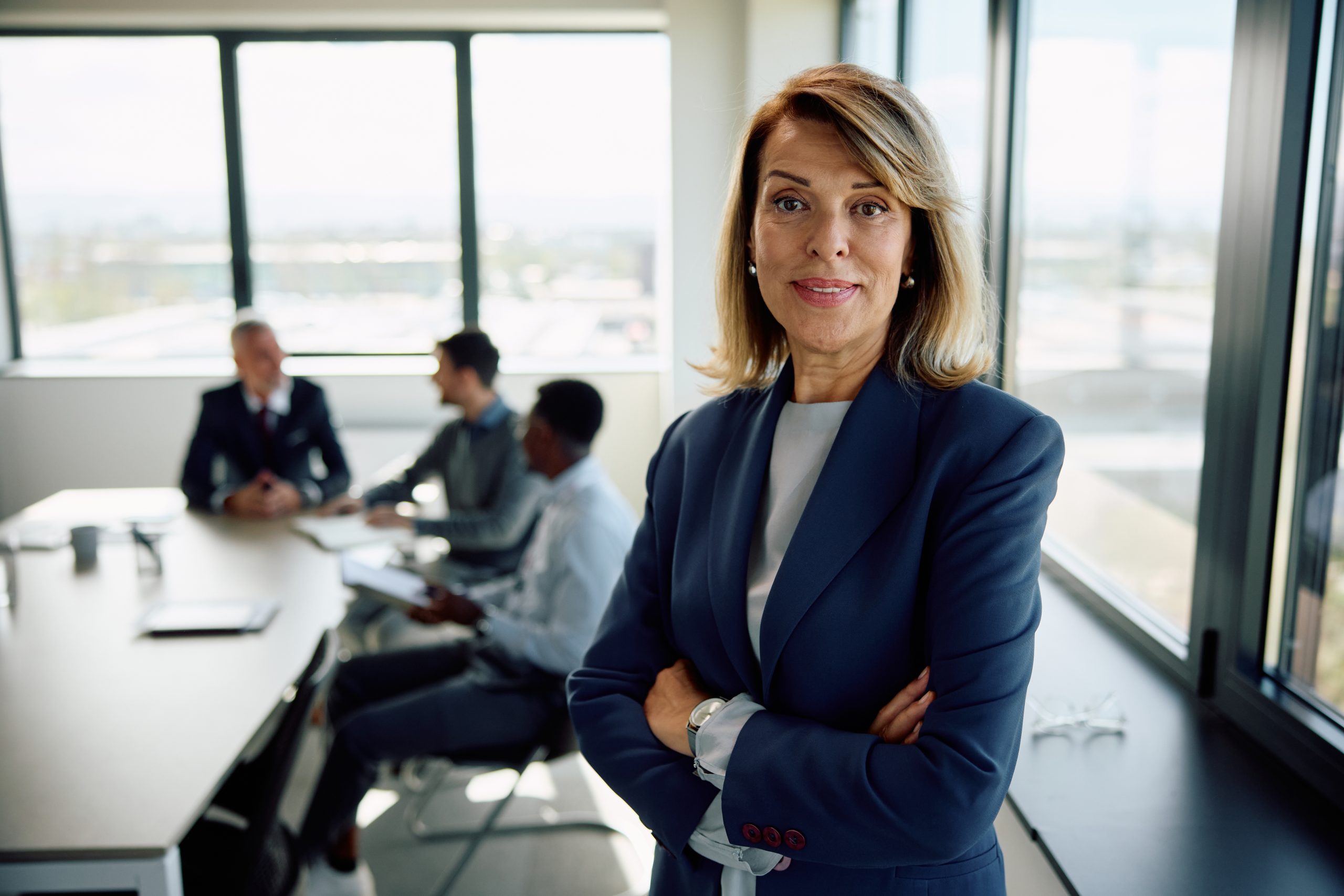 A confident woman in a blue suit stands with arms crossed in an office, while three people have a meeting at a table in the background.