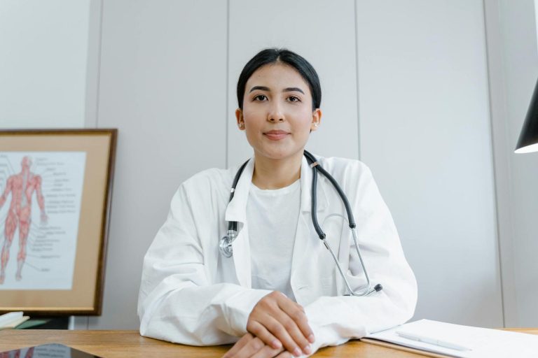 A doctor wearing a white coat and stethoscope sits at a desk, facing the camera. A medical chart of a human body is visible in the background.