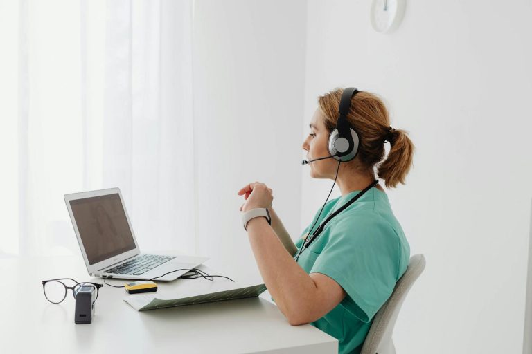 A healthcare worker in scrubs and a headset sits at a desk, speaking to someone on a laptop during a video call. Glasses and medical devices are on the desk.
