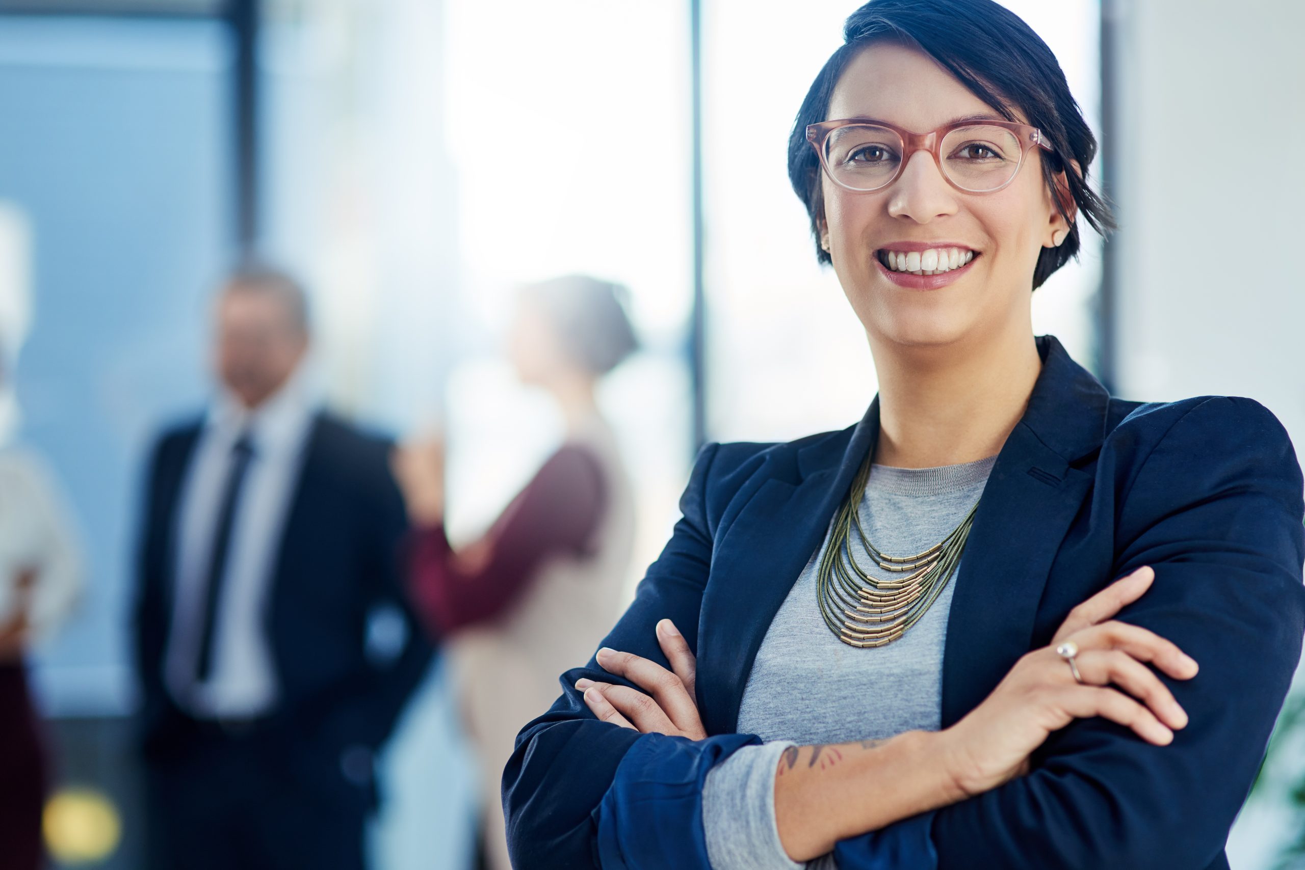 Smiling person with short dark hair and glasses stands with arms crossed; three blurred people are talking in the background in an office setting.