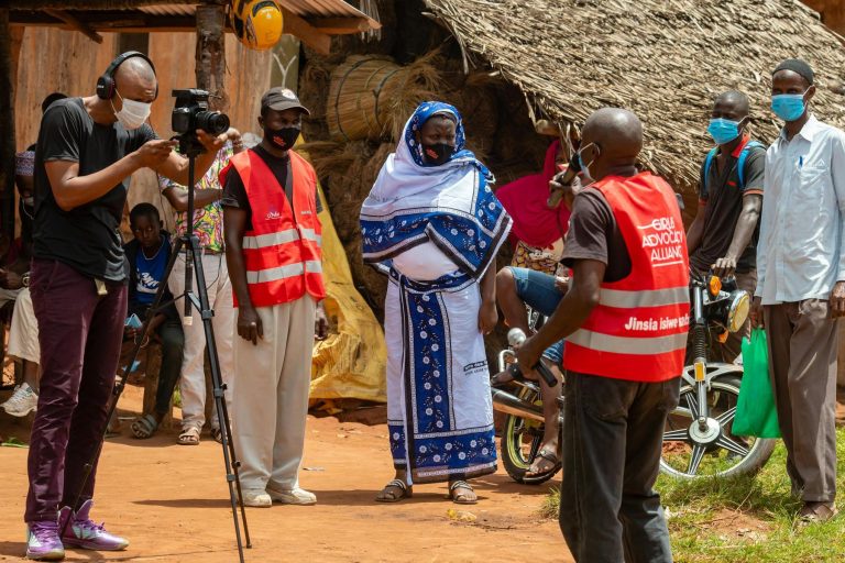 A group of people, some wearing masks and red vests, stand outdoors while one person films with a camera on a tripod in a rural setting.