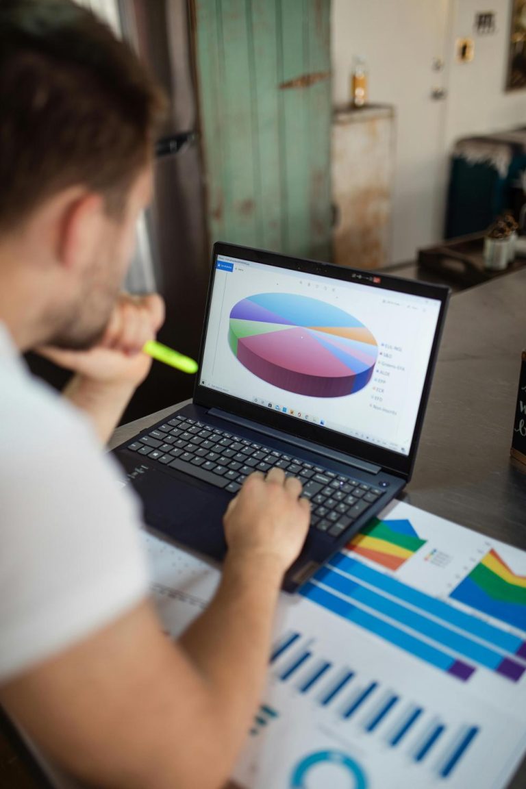 A person looks at a laptop displaying a colorful 3D pie chart, with printed bar and line graphs spread out on the table nearby.