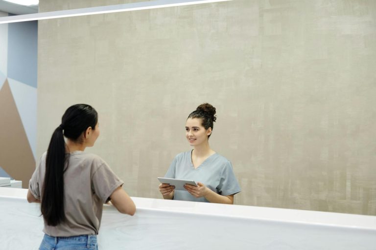 A woman stands at a reception desk while a receptionist in scrubs holds a tablet and speaks to her.