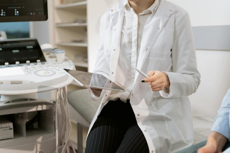 A medical professional in a white coat holds a tablet and stylus in an exam room, sitting next to an ultrasound machine.