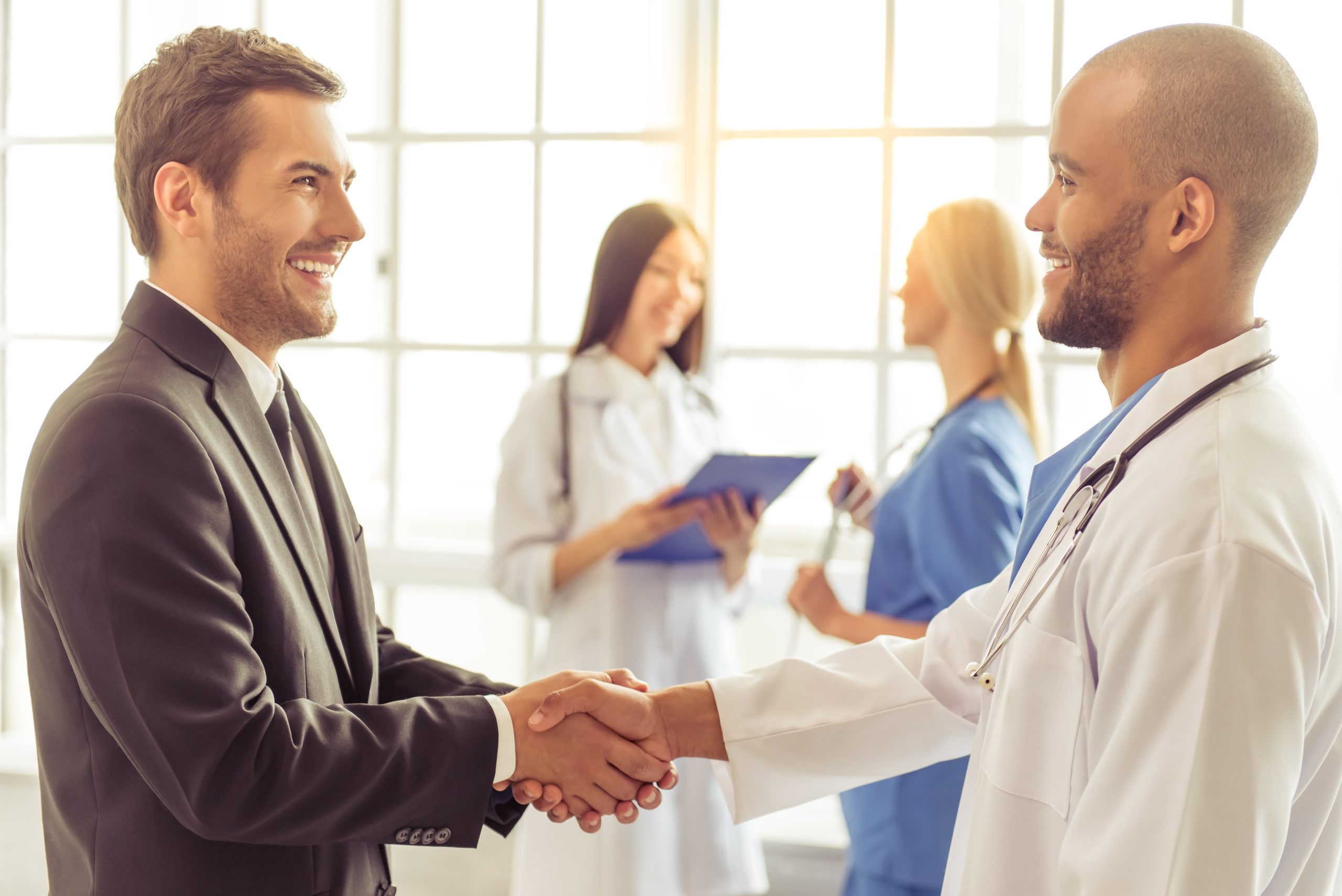 A man in a suit and a doctor shake hands, possibly finalizing an agreement to sell medical practice, while two other medical professionals talk and take notes in the background.