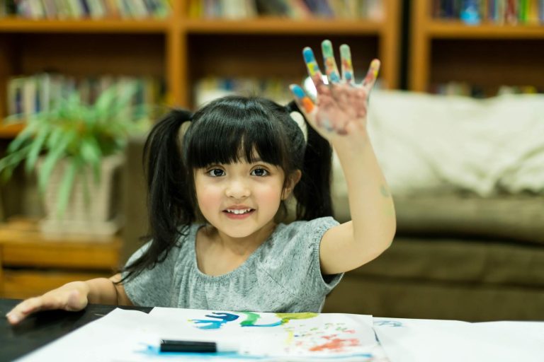 A young girl with pigtails raises her paint-covered hand while sitting at a table with paper and art supplies—a lively scene reflecting the creativity nurtured in Australia's 2025 Early Learning Investment environments. Bookshelves and a couch are in the background.