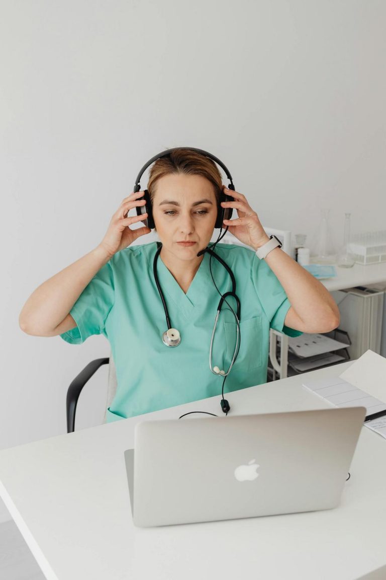A healthcare professional in green scrubs with a stethoscope adjusts headphones while sitting at a desk in front of an open laptop.