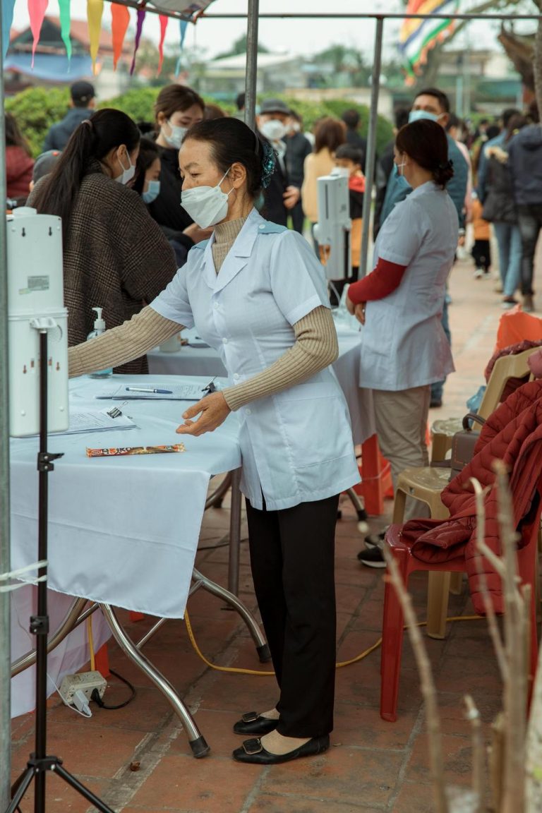 Medical staff wearing masks work at an outdoor registration table, with people standing and waiting in line under a tent.