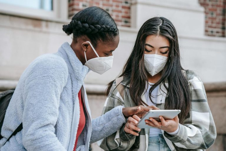 Two young women wearing face masks look at and interact with a tablet device outdoors, one pointing at the screen.