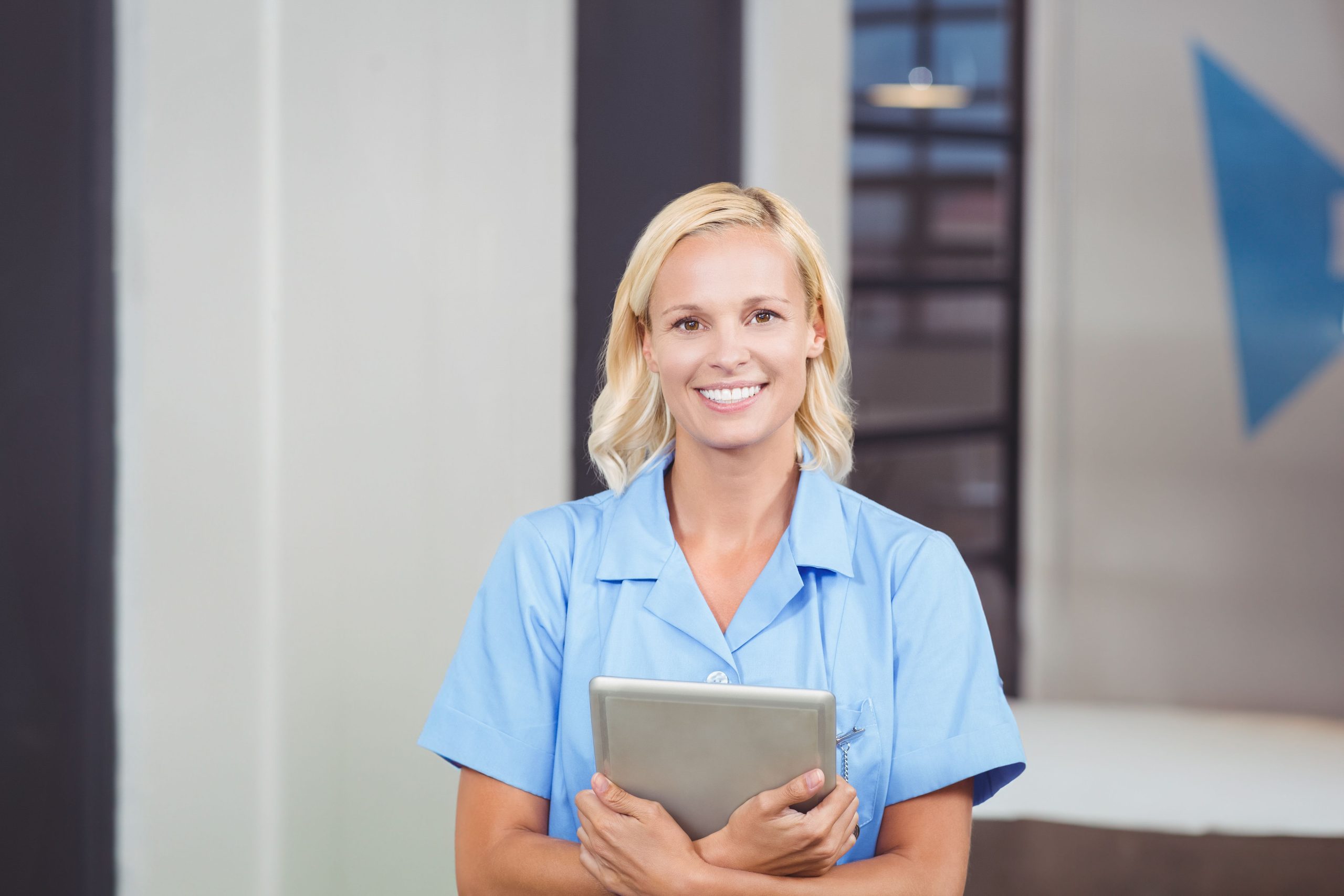 A woman in light blue scrubs, likely a GP practice manager, holds a tablet and smiles at the camera, standing indoors in a professional setting.