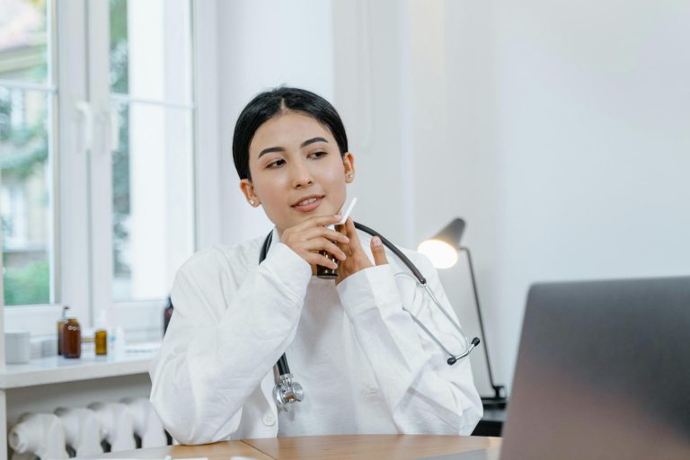 A doctor with a stethoscope around her neck sits at a desk, holding a cup and looking at a laptop in a bright office.