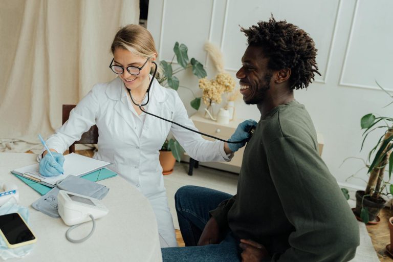 A doctor in a white coat uses a stethoscope to listen to a patient's chest while taking notes at a desk in a medical office.