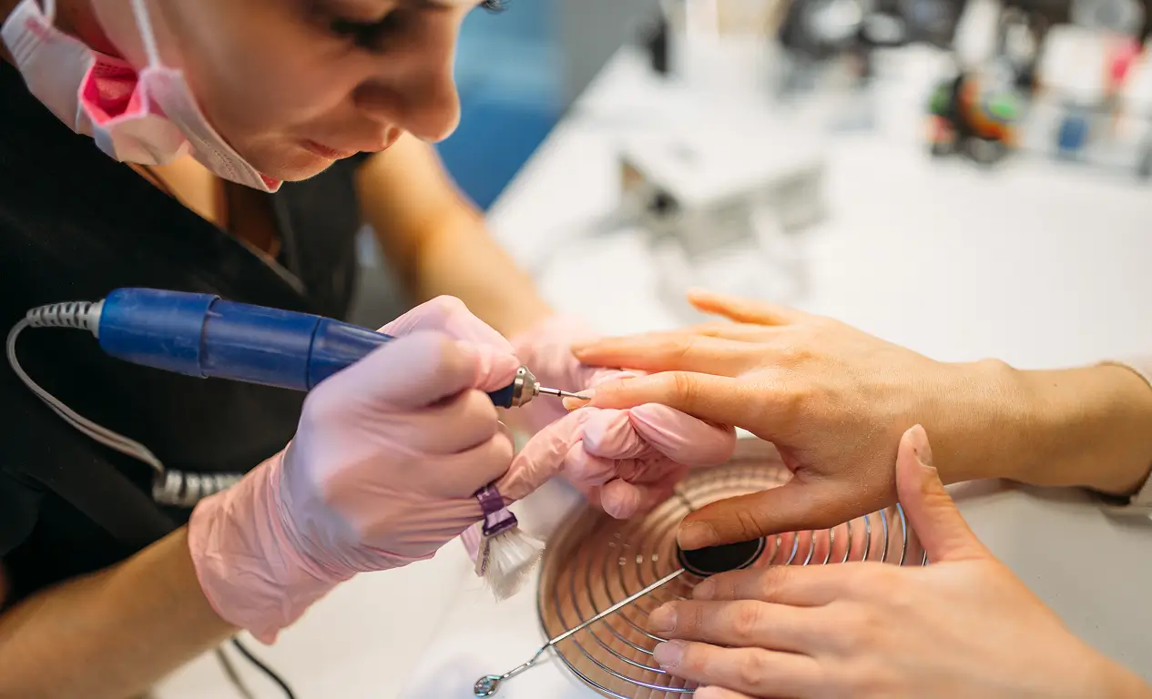 A nail technician wearing pink gloves uses an electric nail file on a client’s fingernails at a manicure station.
