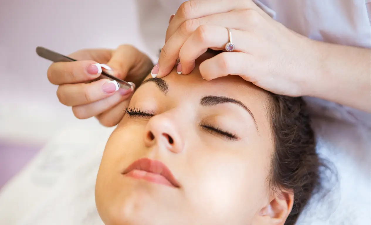 A person with closed eyes is having their eyebrows shaped with tweezers by another person in a close-up view.